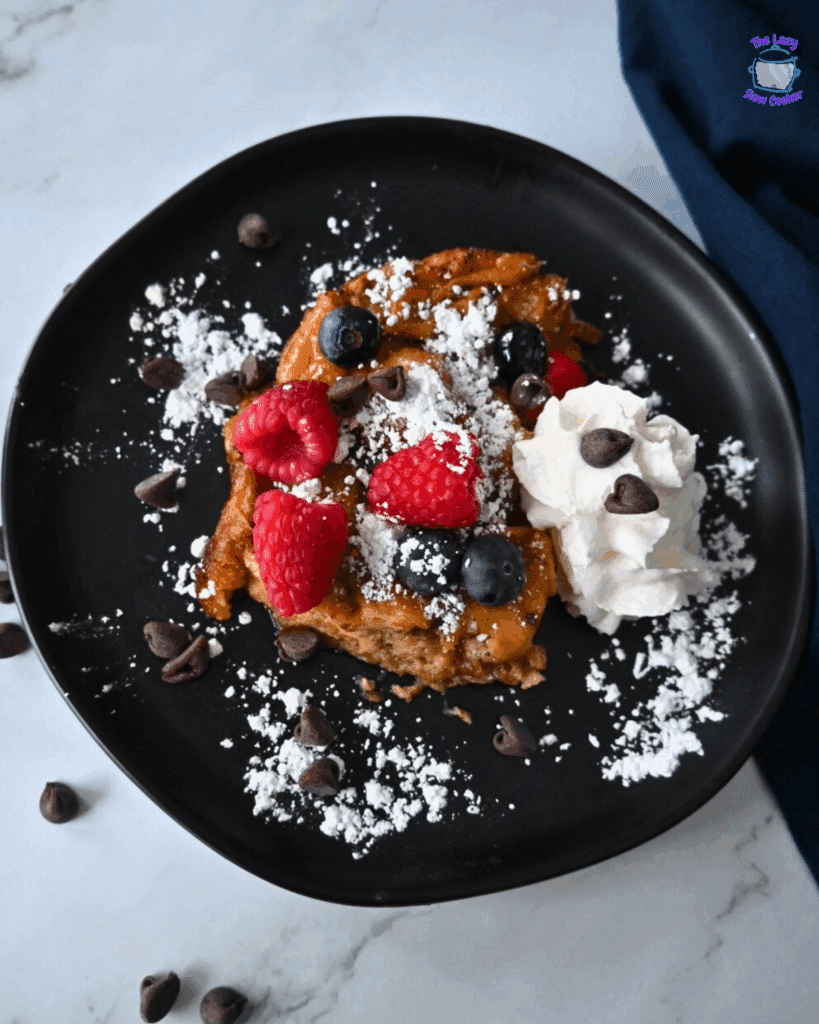 A black plate with a serving of slow cooker donut casserole topped with powdered sugar, raspberries, blueberries, chocolate chips, and a dollop of whipped cream, on a marble surface.
