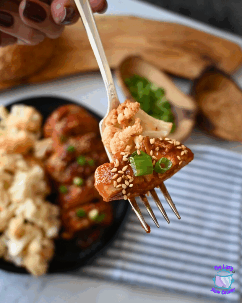 A fork holding a piece of saucy slow cooker chicken topped with sesame seeds, green onions, and cauliflower. In the background is a black plate with more tofu, cauliflower, and chopped green onions.