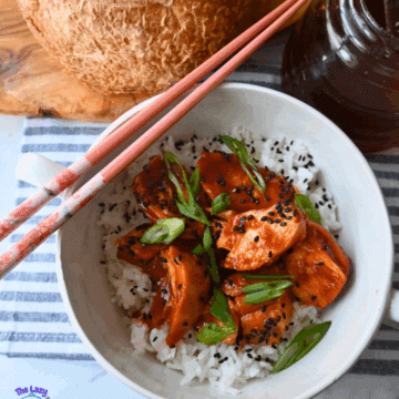 A white bowl of rice topped with slow cooked honey garlic chicken, black sesame seeds, and sliced green onions. Pink chopsticks rest on the bowl. A round loaf of bread and a glass jar of dark sauce are nearby on a striped cloth.