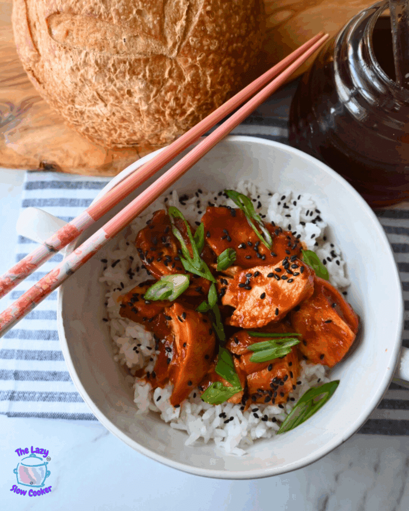 A white bowl of rice topped with slow cooked honey garlic chicken, black sesame seeds, and sliced green onions. Pink chopsticks rest on the bowl. A round loaf of bread and a glass jar of dark sauce are nearby on a striped cloth.