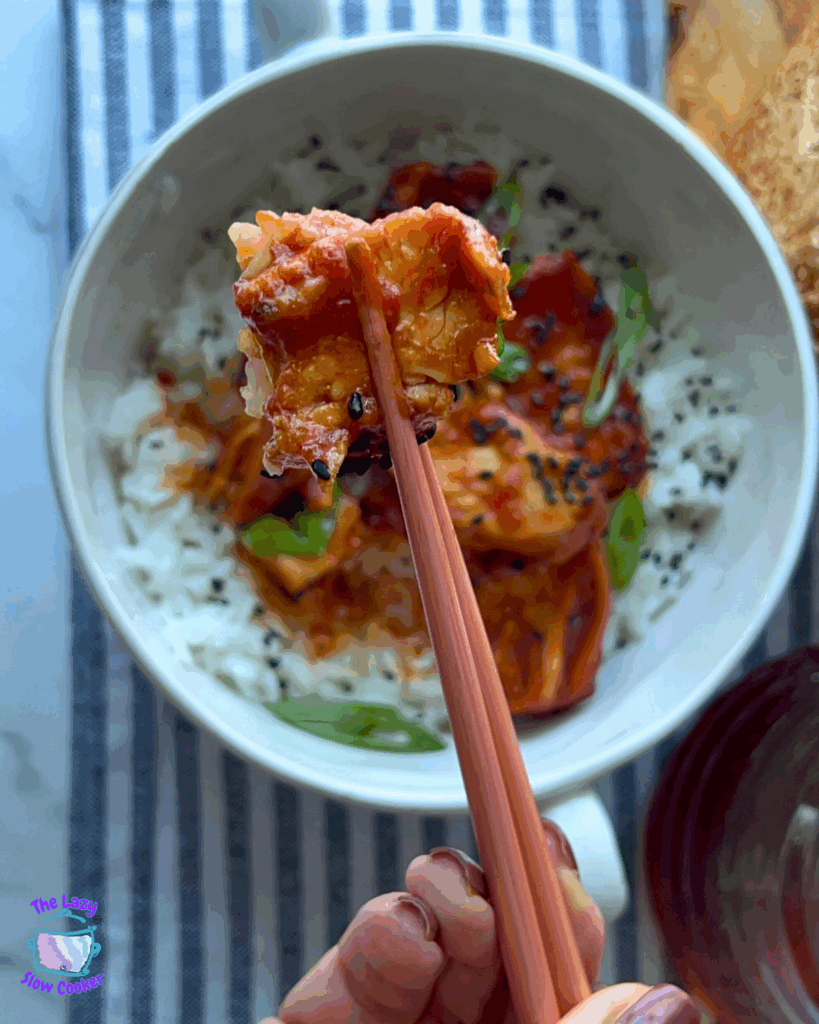 A hand holds chopsticks with a bite of crockpot garlic honey chicken above a bowl of white rice topped with chicken, sauce, green onions, and black sesame seeds. A striped cloth is underneath the bowl.