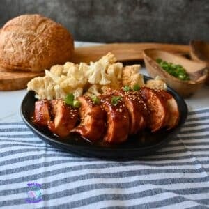 A black plate holds sliced, slow cooker honey garlic chicken breast topped with sesame seeds and chopped green onions, alongside steamed cauliflower florets. A round loaf of bread and a wooden bowl of green onions are nearby.