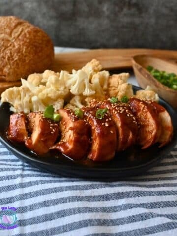 A black plate holds sliced, slow cooker honey garlic chicken breast topped with sesame seeds and chopped green onions, alongside steamed cauliflower florets. A round loaf of bread and a wooden bowl of green onions are nearby.