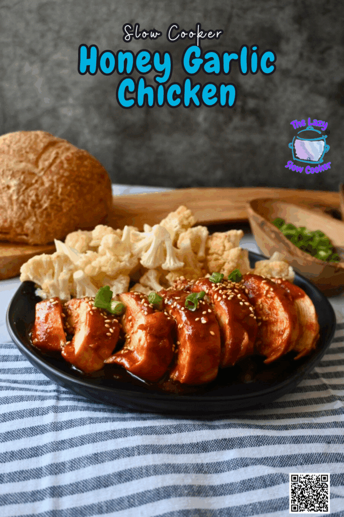 A black plate holds sliced, crockpot honey garlic chicken breast topped with sesame seeds and chopped green onions, alongside steamed cauliflower florets. A round loaf of bread and a wooden bowl of green onions are nearby.