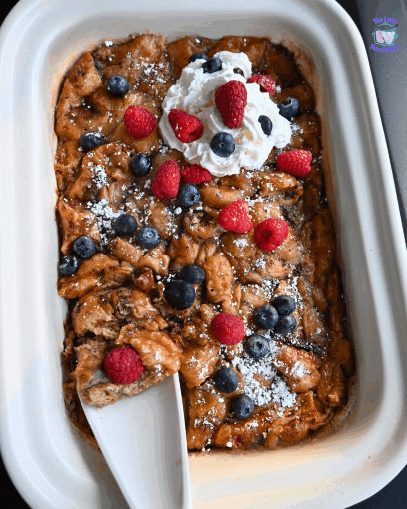 A slow cooker casserole dish filled with golden-brown donut bread pudding, topped with a generous dusting of powdered sugar, whipped cream, and fresh berries. A white spoon is about to scoop some out.