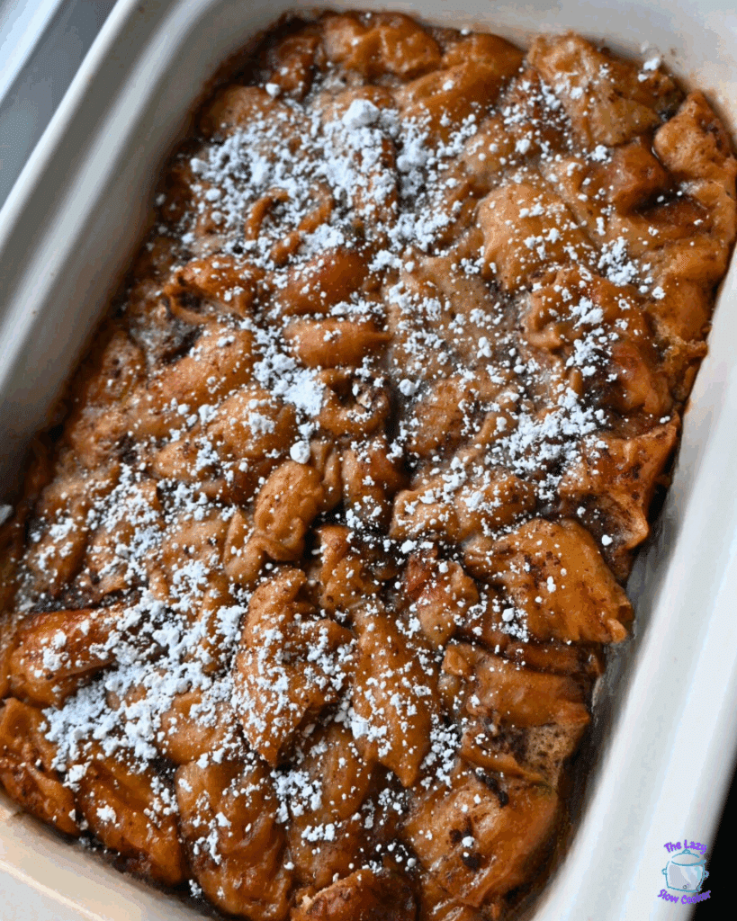 A slow cooker casserole dish filled with golden-brown bread pudding, topped with a generous dusting of powdered sugar.