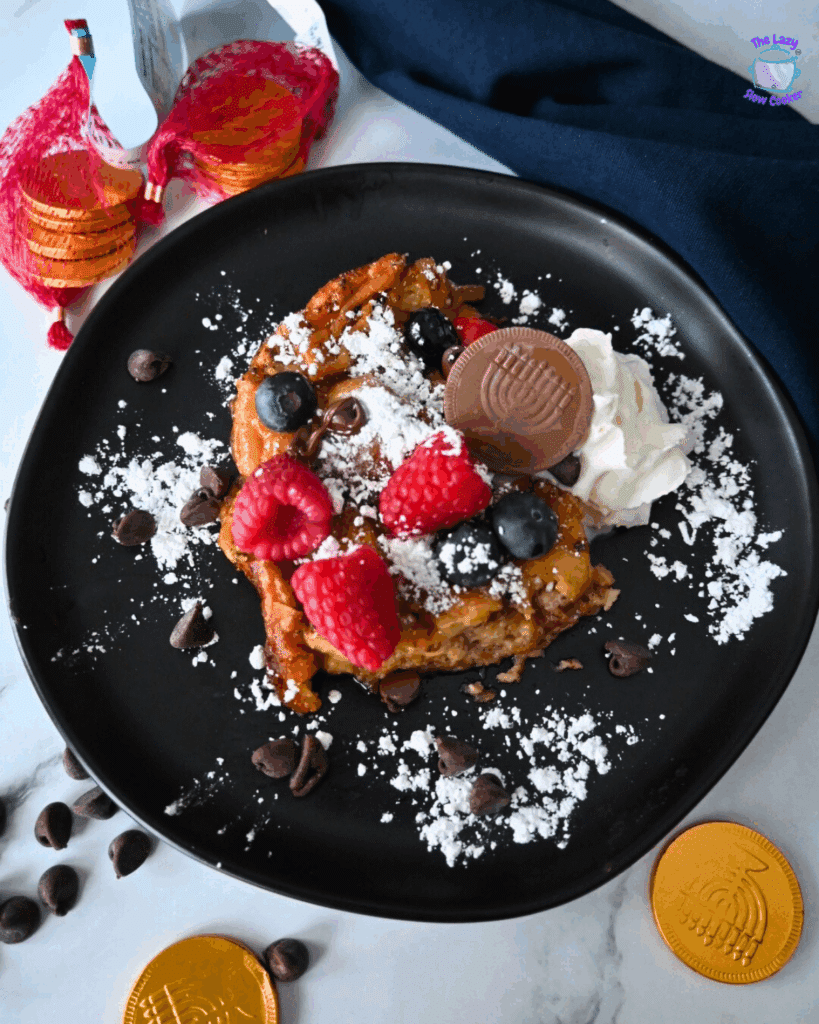 A black plate with a serving of donut casserole topped with powdered sugar, raspberries, blueberries, chocolate chips, and a dollop of whipped cream, on a marble surface.