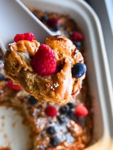 A close-up of a serving of donut bread pudding topped with fresh raspberries and blueberries being lifted from a slow cooker. The dish is golden brown with a dusting of powdered sugar.