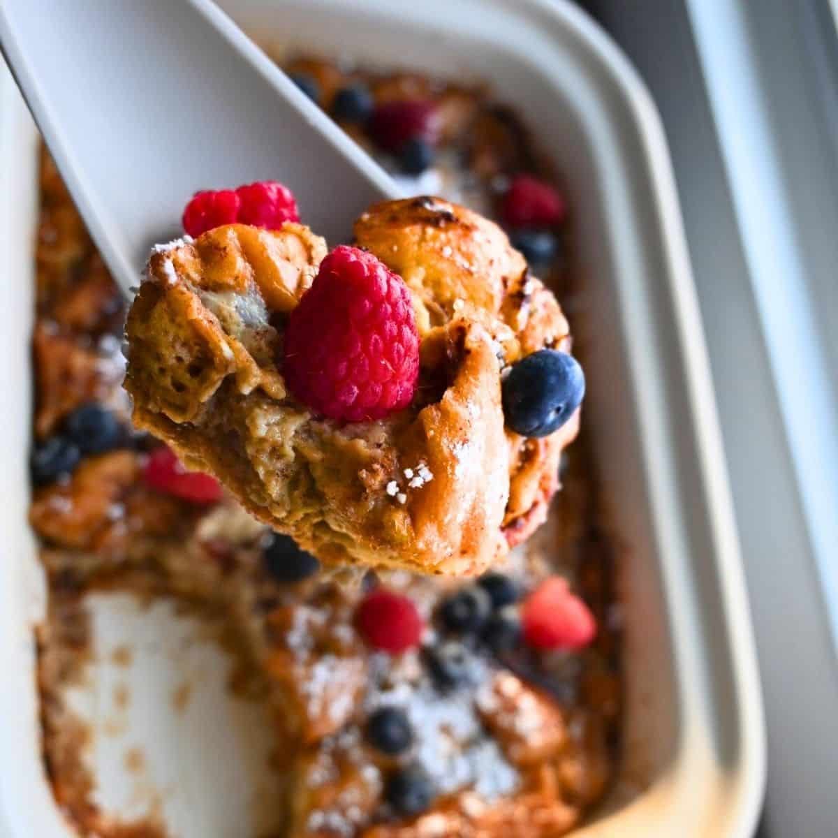 A close-up of a serving of donut bread pudding topped with fresh raspberries and blueberries being lifted from a slow cooker. The dish is golden brown with a dusting of powdered sugar.