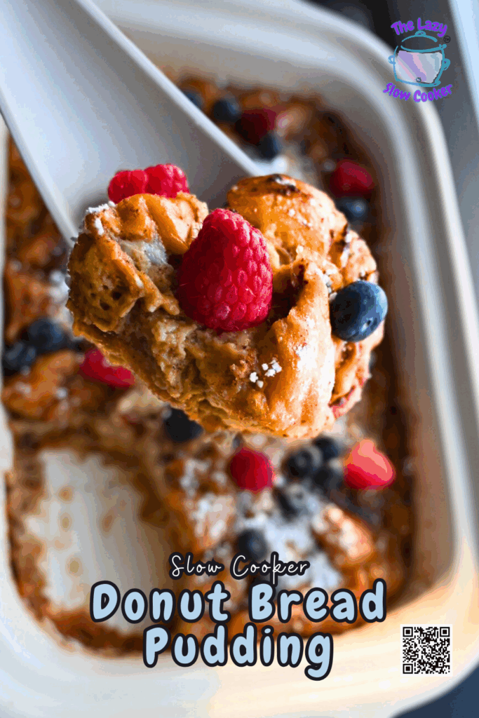 A close-up of a serving of donut bread pudding topped with fresh raspberries and blueberries being lifted from a slow cooker. The dish is golden brown with a dusting of powdered sugar. Text reads: Slow Cooker Donut Bread Pudding.