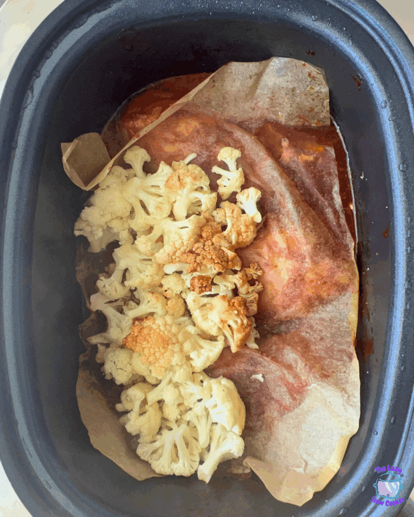 A slow cooker with parchment paper lining, containing roasted cauliflower florets on top of a layer of sauce and cooked meat.