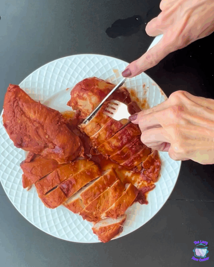 A person uses a fork and knife to cut into a piece of sliced, cooked, saucy chicken breast on a white textured plate, set on a dark surface.