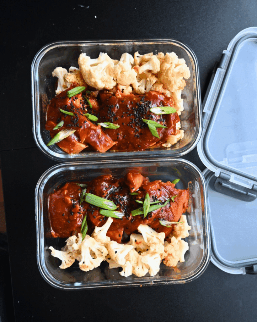Two glass meal prep containers filled with rice, roasted cauliflower, and pieces of honey garlic chicken in a reddish sauce, garnished with chopped green onions and black sesame seeds, with one containers lid resting nearby.