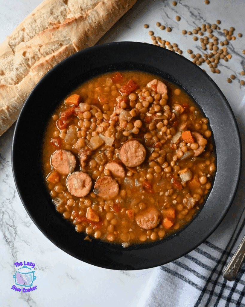 A black bowl filled with slow cooker lentil soup containing sausage slices, carrots, and tomatoes, placed on a striped towel. A baguette and a spoon are next to the bowl on a marble surface.
