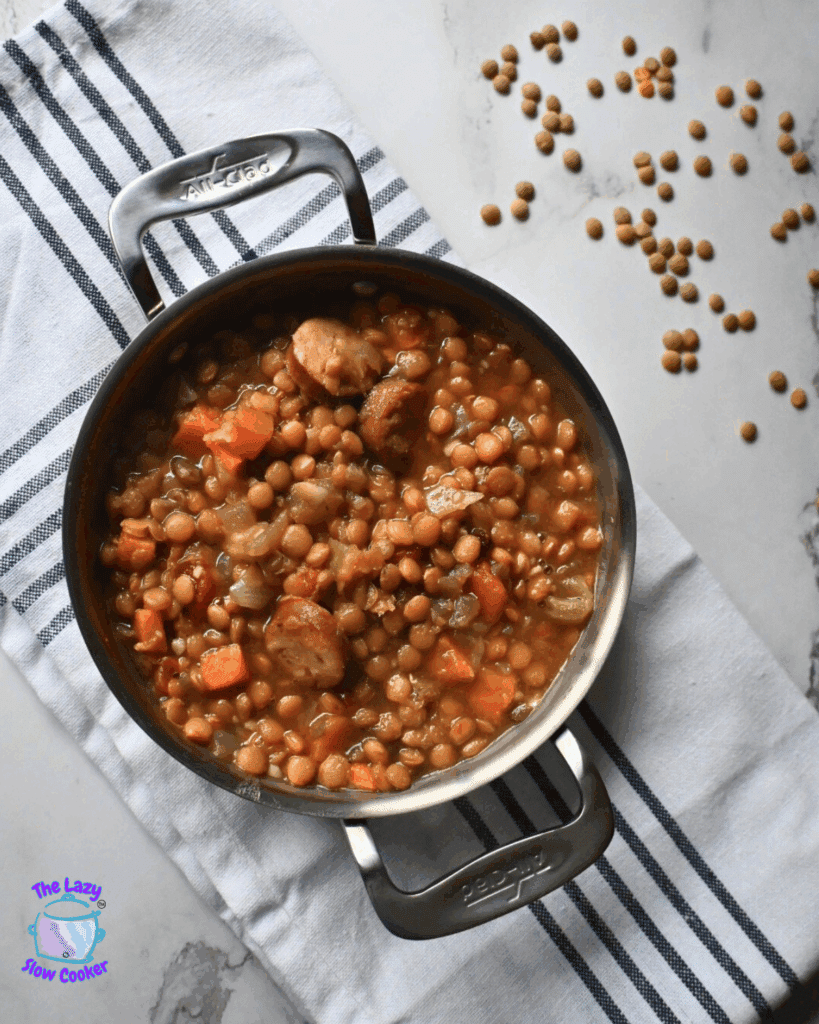 Overhead view of a handled serving plate filled with crockpot lentil stew containing sausage, carrots, and onions, resting on a striped kitchen towel. Uncooked lentils are scattered on the white countertop nearby.