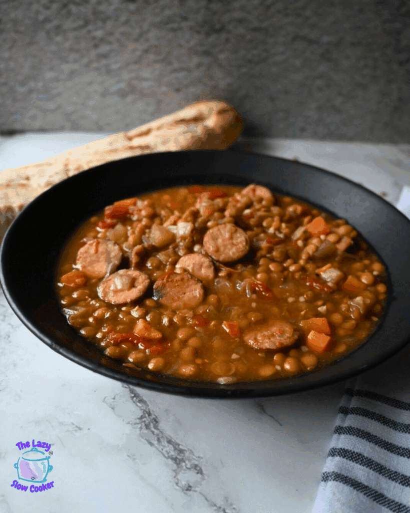 A black bowl filled with lentil soup and sliced sausage sits on a marble surface, with a piece of bread in the background and a striped cloth to the side.