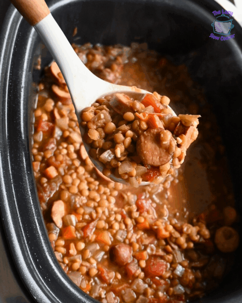 A white spoon holds up a serving of lentil stew with sliced sausage, carrots, celery, and tomatoes over a slow cooker filled with the same hearty mixture.