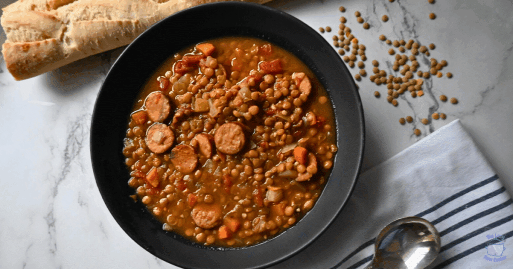 A black bowl filled with slow cooker lentil soup containing sausage slices, carrots, and tomatoes, placed on a striped towel. A baguette and a spoon are next to the bowl on a marble surface.