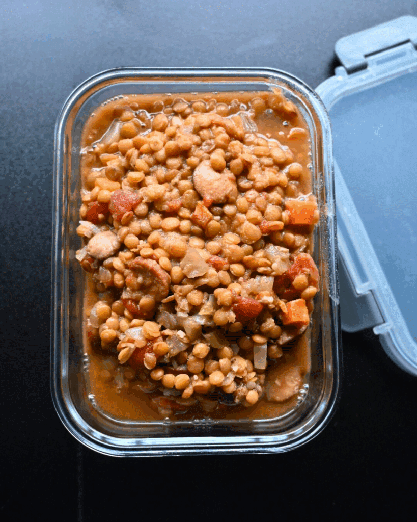 A glass container filled with leftover slow cooker sausage and lentil stew containing chopped carrots, tomatoes, onions, and sausage slices, with its plastic lid open on a dark surface.