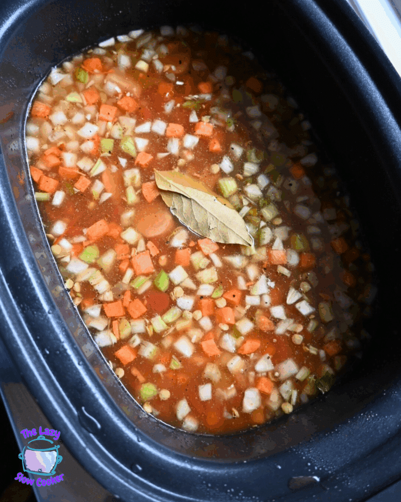A slow cooker filled with diced carrots, celery, onions, and tomatoes in broth, topped with a bay leaf, ready to cook.