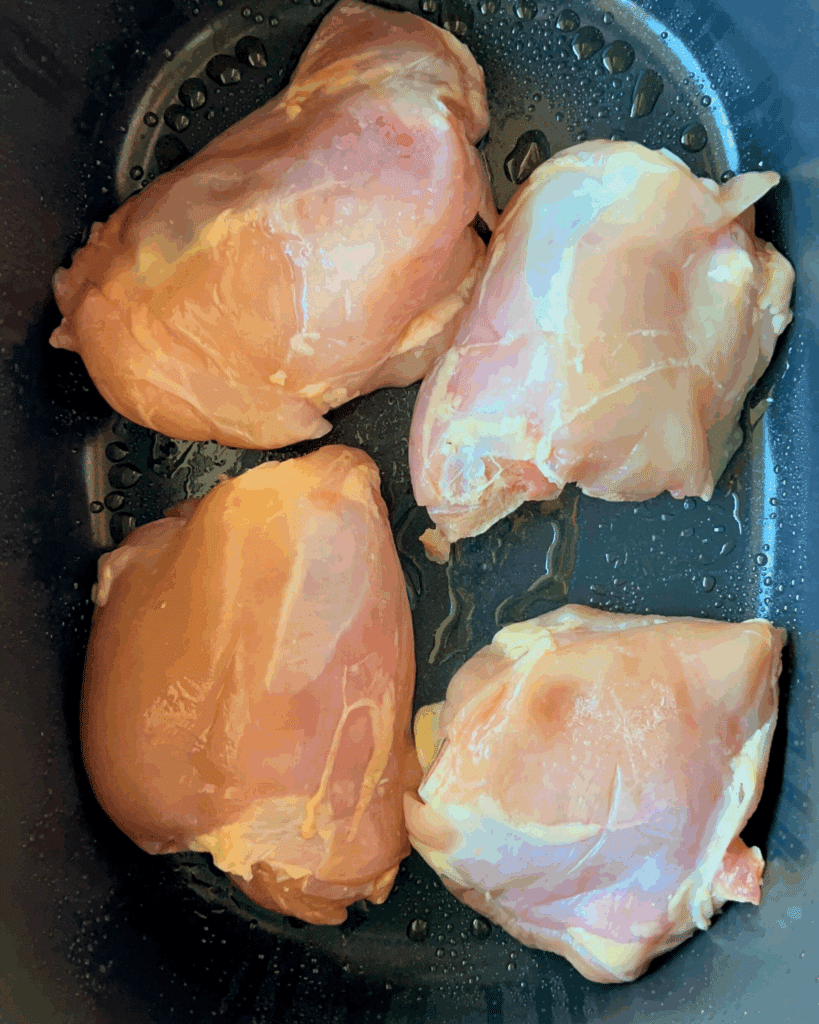 Four raw boneless, skinless chicken breasts placed in a dark, shallow container with droplets of water or oil visible around them.