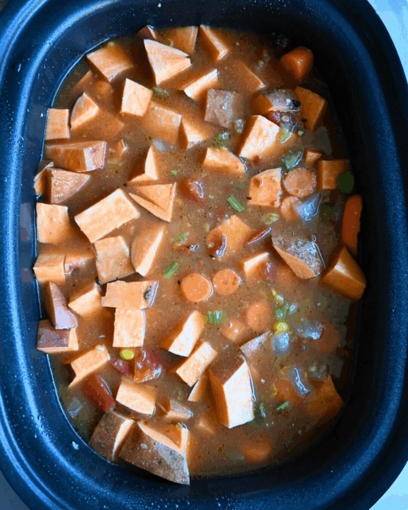 Chunks of sweet potatoes, carrots, and green onions in a reddish-brown broth inside a black slow cooker or casserole dish, ready to be cooked.