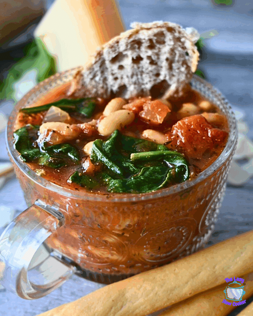 A clear soup mug filled with slow cooked tomato and white bean soup garnished with spinach and a slice of bread.