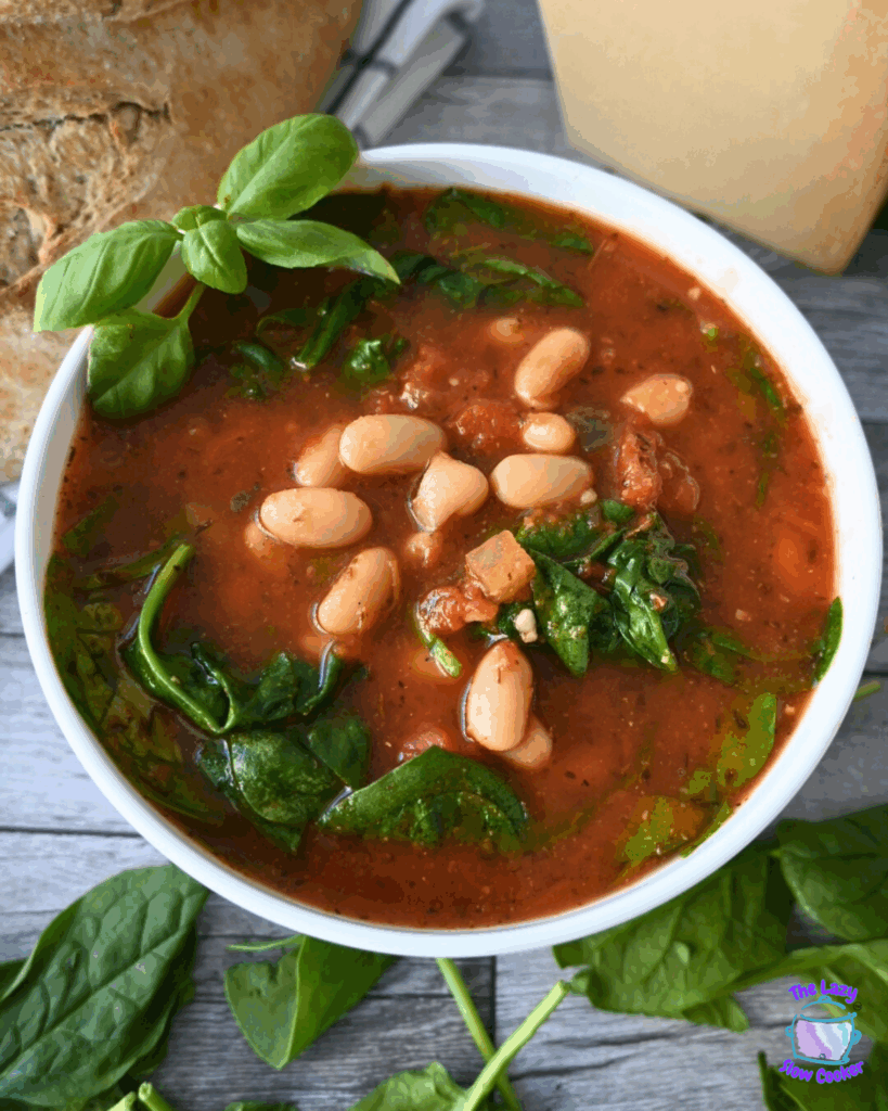 A bowl of tomato and white bean soup with spinach, garnished with fresh basil. The text reads Slow Cooker Tomato & White Bean Soup. A QR code and a logo are in the corners. The bowl is on a wooden surface.
