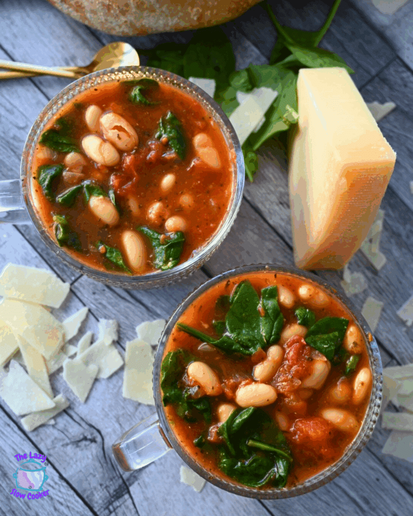 Two clear glass soup mugs filled with tomato-based soup containing white beans and spinach, surrounded by shaved parmesan cheese, a block of parmesan, and fresh spinach on a wooden surface.