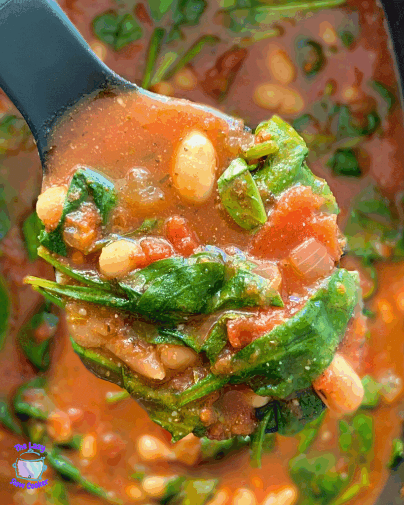 A close-up of a ladle scooping a chunky tomato-based soup with white beans and fresh spinach leaves. The background shows the rest of the soup in the crockpot.