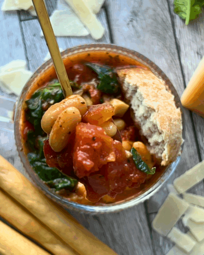 A clear soup mug filled with slow cooked tomato and white bean soup garnished with spinach and a slice of bread.
