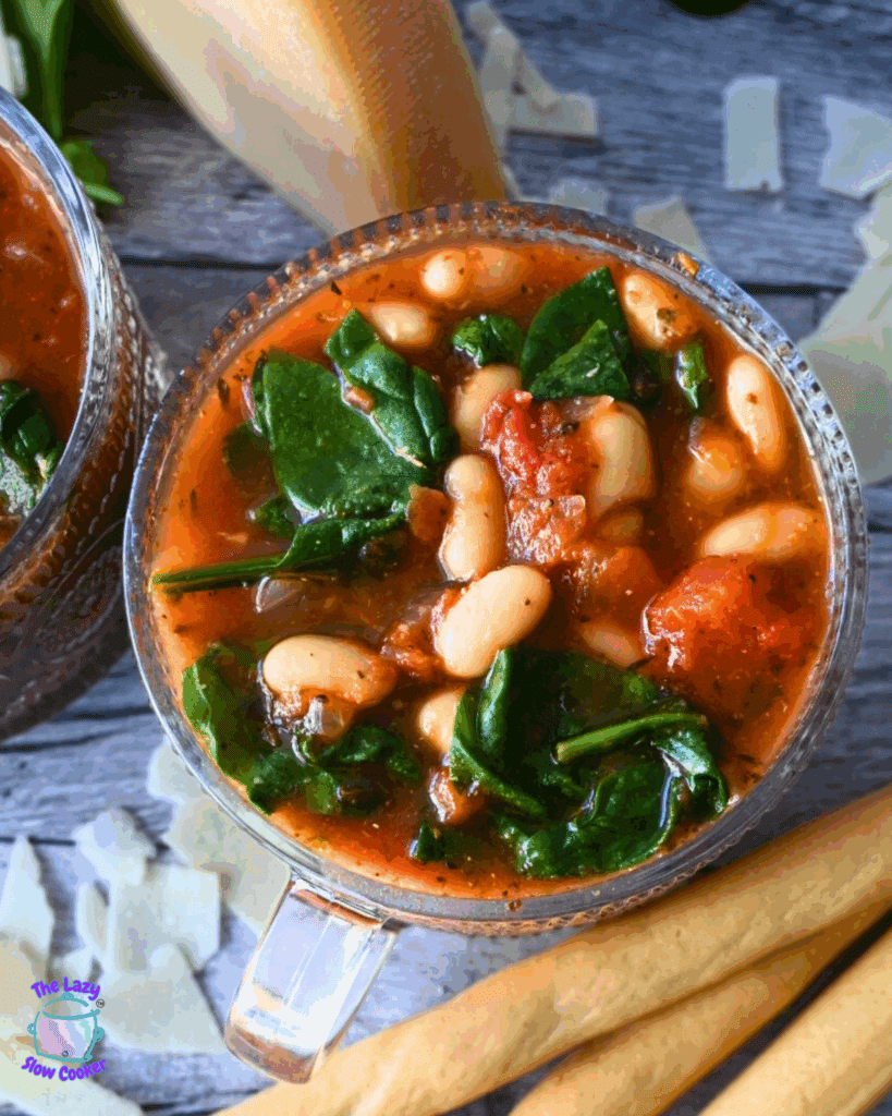A clear soup mug filled with slow cooked tomato and white bean soup garnished with spinach and a slice of bread.