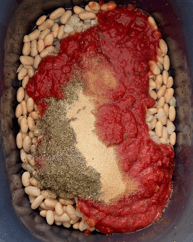An overhead view of white beans topped with tomato sauce, dried herbs, spices, and seasonings in a slow cooker before being mixed together.