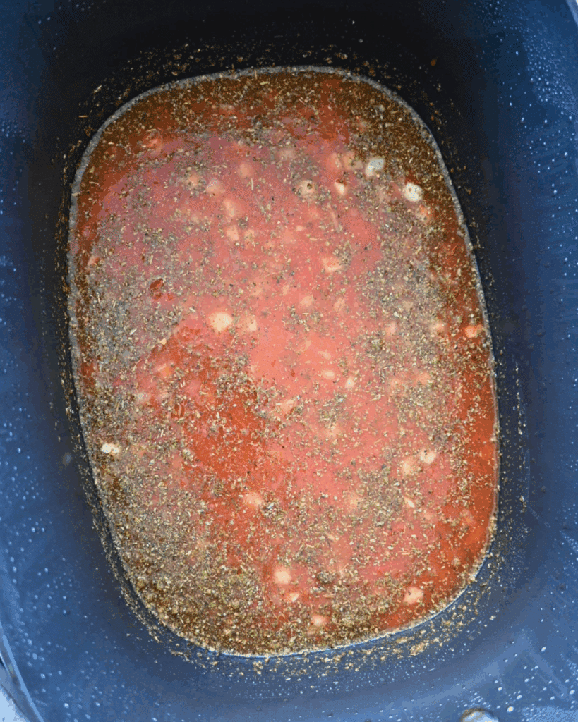 A slow cooker filled with tomato based soup, herbs, spices, and white beans ready for cooking. The sauce has a red color with green and white specks throughout.
