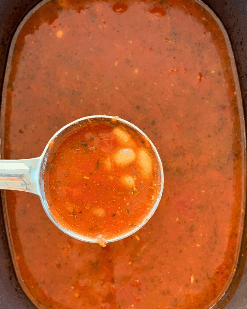 A metal ladle holds up a serving of tomato-based soup with white beans and herbs over a slow cooker filled with the same soup.