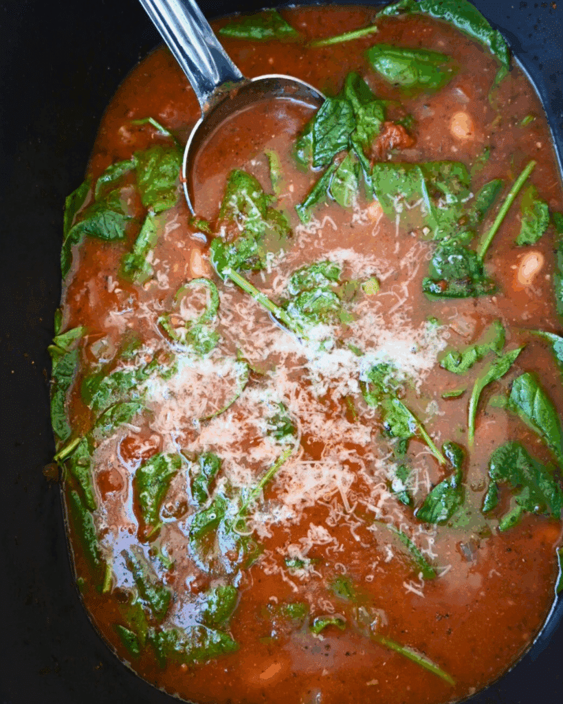 A ladle in a pot of tomato-based soup with fresh spinach leaves and grated cheese sprinkled on top.