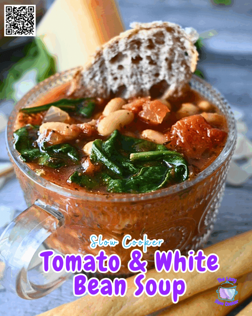 A clear soup mug filled with slow cooked tomato and white bean soup garnished with spinach and a slice of bread. The text “Slow Cooker Tomato & White Bean Soup” is displayed at the bottom in purple and white.