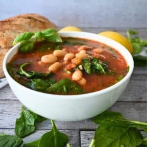 A bowl of tomato and white bean soup with spinach, garnished with fresh basil. The text reads Slow Cooker Tomato & White Bean Soup. A QR code and a logo are in the corners. The bowl is on a wooden surface.