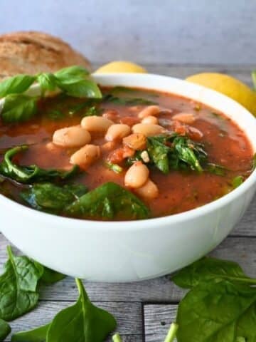 A bowl of tomato and white bean soup with spinach, garnished with fresh basil. The text reads Slow Cooker Tomato & White Bean Soup. A QR code and a logo are in the corners. The bowl is on a wooden surface.