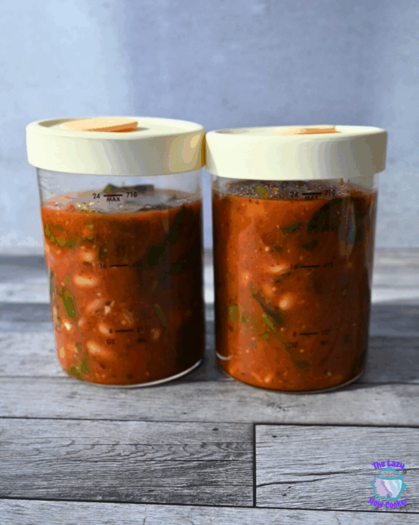 Two glass jars with white lids filled with a chunky red soup containing white beans and green herbs, sitting on a wooden surface with a gray background.