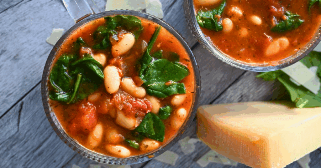 Two bowls of tomato-based soup with white beans and spinach are on a wooden surface, next to a wedge of cheese and spinach leaves.