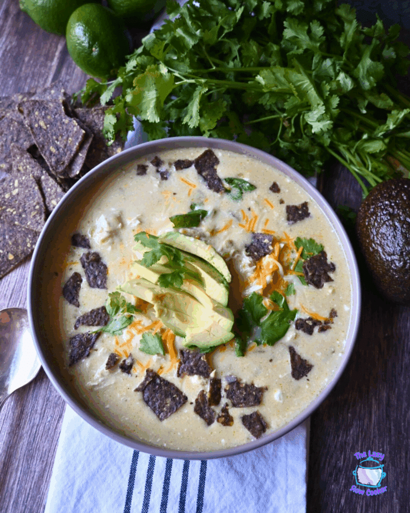 A bowl of creamy soup topped with sliced avocado, shredded cheese, cilantro, and crumbled blue corn tortilla chips, with fresh cilantro, limes, an avocado, and blue corn chips on the side.