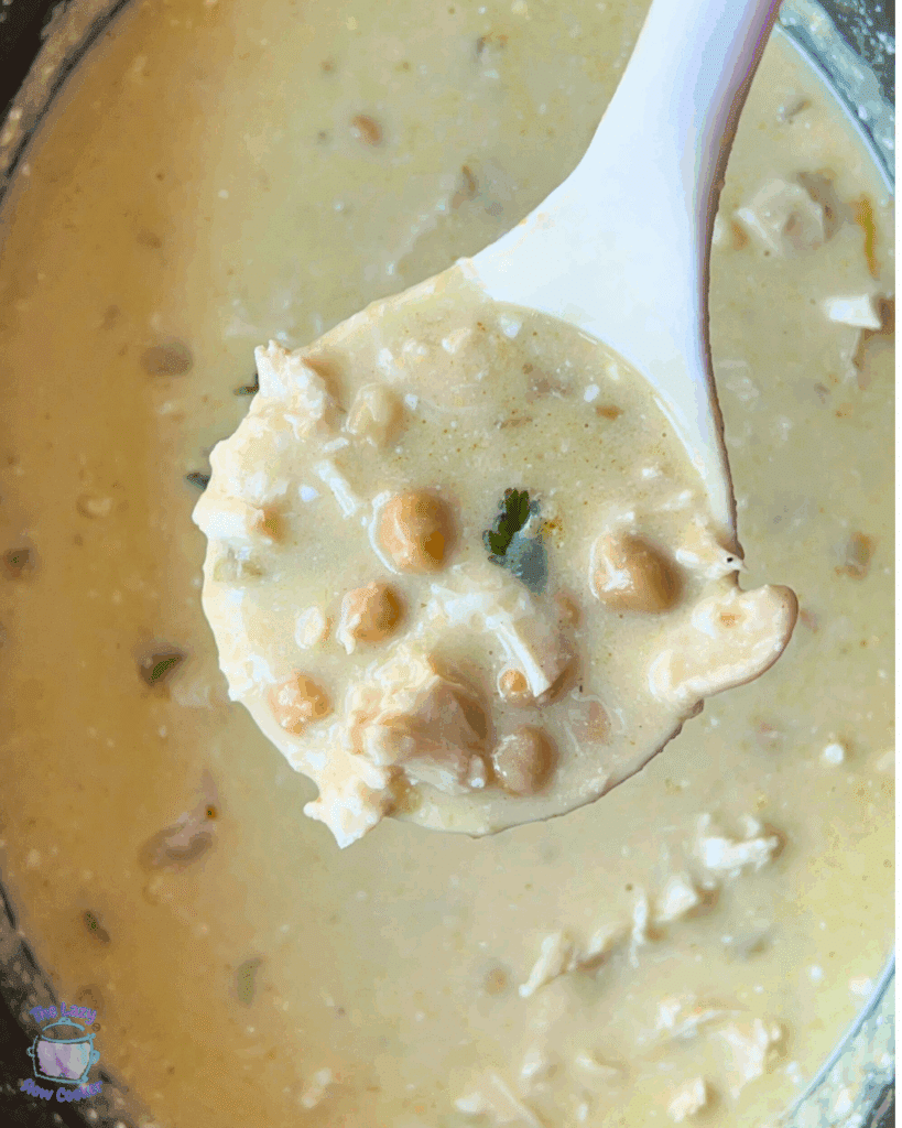 A close-up of a white ladle holding creamy enchilada chicken soup with chickpeas, shredded chicken, herbs, and visible broth, over a pot filled with the same soup.