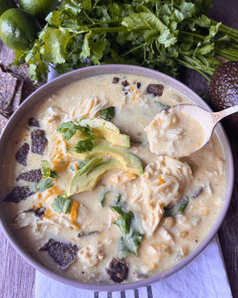 A bowl of creamy chicken soup topped with avocado slices, shredded cheese, cilantro, and tortilla chips. A spoon is scooping some soup. Fresh cilantro, lime, and an avocado are in the background.