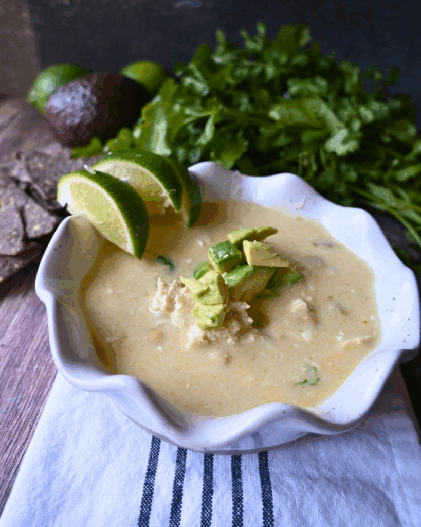 A bowl of creamy enchilada chicken soup topped with diced avocado, with lime wedges on the rim. Fresh cilantro is in the background, and the bowl sits on a white cloth with dark stripes.