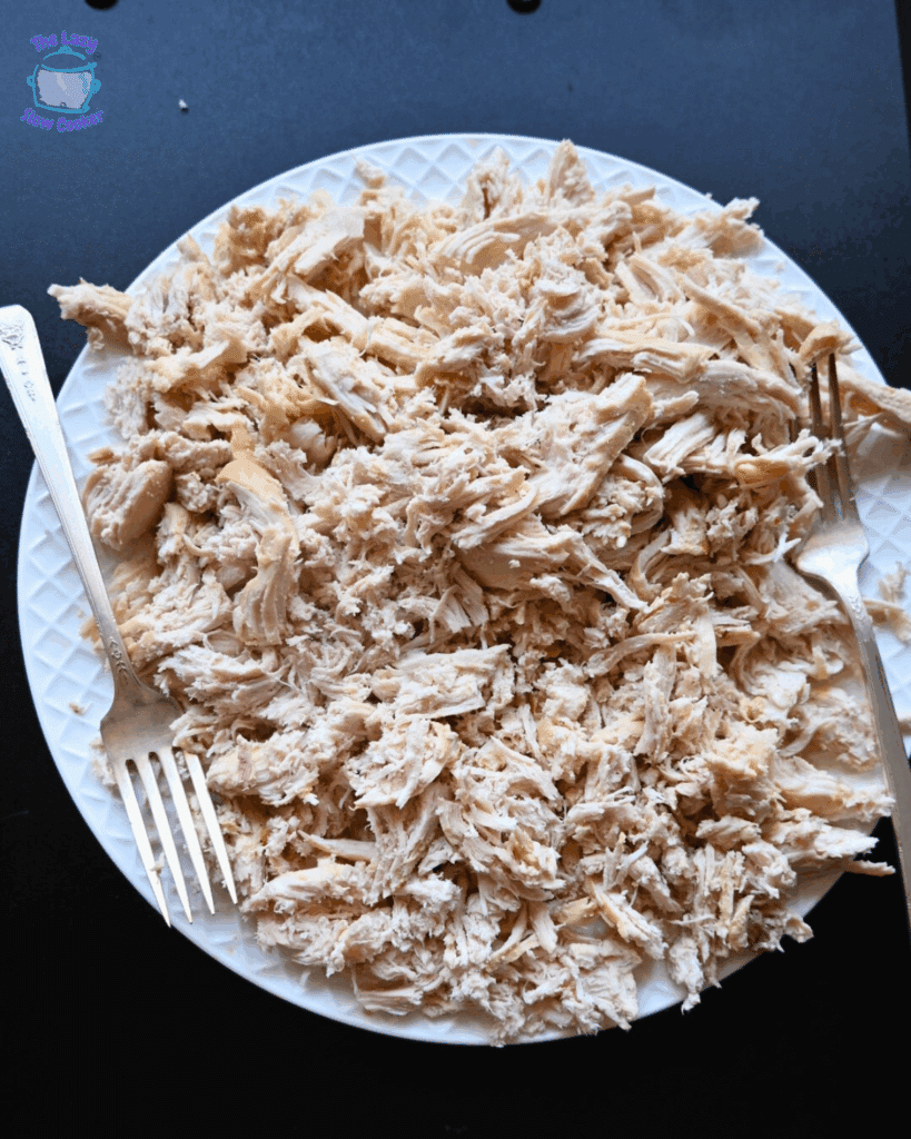A white plate filled with shredded cooked chicken sits on a dark surface. Two forks rest on the plate, used for shredding the chicken.