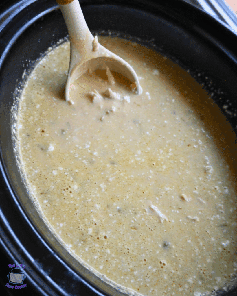 A slow cooker filled with creamy enchilada chicken soup, being stirred with a beige ladle. The soup appears thick with small chunks visible throughout.