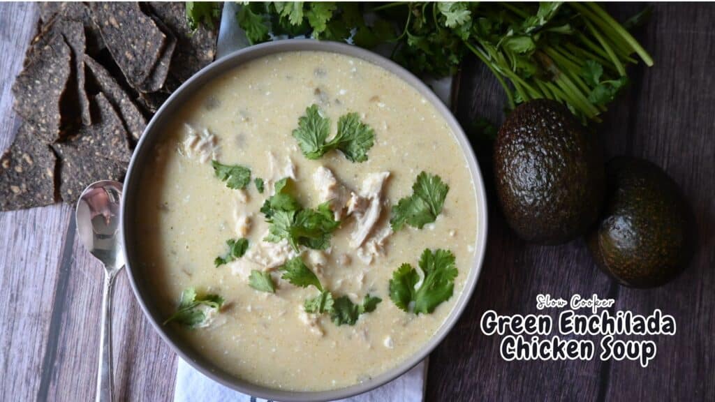 A bowl of creamy green enchilada chicken soup topped with cilantro sits on a wooden table next to blue corn tortilla chips, fresh cilantro, and two avocados. Text on image reads: Slow Cooker Green Enchilada Chicken Soup.