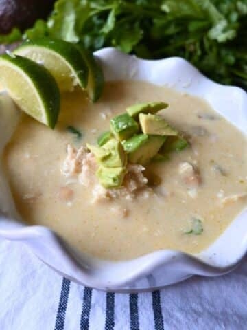 A bowl of creamy enchilada chicken soup topped with diced avocado, with lime wedges on the rim. Fresh cilantro is in the background, and the bowl sits on a white cloth with dark stripes.