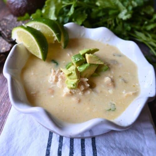 A bowl of creamy enchilada chicken soup topped with diced avocado, with lime wedges on the rim. Fresh cilantro is in the background, and the bowl sits on a white cloth with dark stripes.